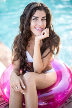 Cheerful girl in a white swimsuit sits on the pink rubber ring and looks into the camera with a smile on the background of the swimming pool. She holds her left hand under the chin. Vertical.