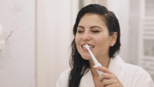 Bathroom Mirror, Lady Brushing, Morning Routine. Attractive mature woman brushes teeth after shower, viewing reflection in bathroom mirror, clad in white silk bathrobe