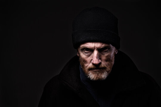 Portrait of a grumpy-looking older, bearded man wearing woollen fisherman's hood, scowling at camera. Dramatic lighting, isolated on black. Ominous, aggressive and dangerous feel. Copy space.