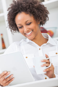 A beautiful happy mixed race African American girl or young woman using a tablet computer in her kitchen and drinking mug of coffee or tea