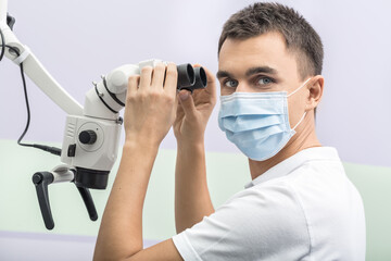Friendly doctor is looking into the camera while holding microscope with his hands in the clinic on the wall background. He wears a white uniform and a blue medical mask. Horizontal.