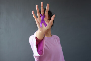 Woman in purple t-shirt holding showing purple awareness ribbon on palm, covering face. Symbol is used to raise awareness for Alzheimer's disease, domestic abuse, cancer disease