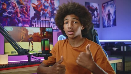 African American teenage gamer, blogger speaks on camera using microphone, shows thumbs up. Recording video games review for social networks. Neon cozy room. PC with online 3D shooter on background.
