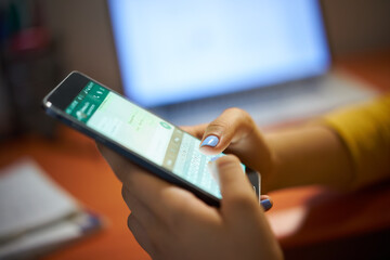 Young woman using cell phone to send text message on social network at night. Closeup of hands with computer laptop in background
