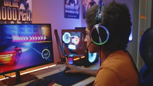 Professional African American teenage gamer in headphones plays in online 3D racing game on powerful PC in cozy room in the evening. Video game live streaming or cybersport championship. Dolly shot.