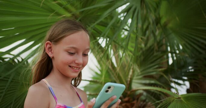 Happy teen girl showing selfie on smartphone under palm tree, leaves background. Resting in tropics on vacations. Blonde smiling teenager photographing herself for social media. Travel, tourism.