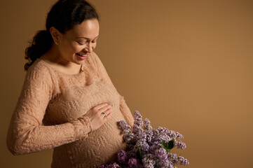 Happy smiling gravid woman, expectant mother strokes her pregnant belly, poses with bouquet of...