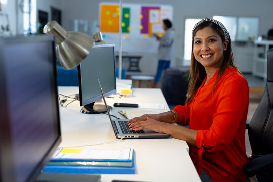 Portrait of happy biracial casual businesswoman using laptop in office