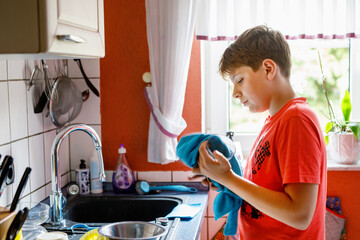 Handsome teenager boy drying a clean plate with a towel. Preteen child helping in kitchen with housework.