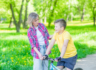 Sporty family leisure. Happy mother talks with her young son, who ride a bike in summer park