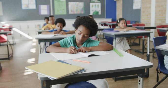 Focused diverse schoolchildren writing at desks in elementary school classroom, slow motion
