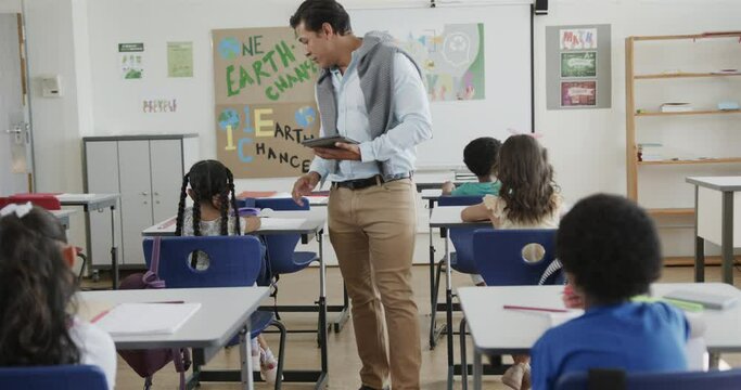 Diverse male teacher with tablet helping children at desks in elementary school class, slow motion