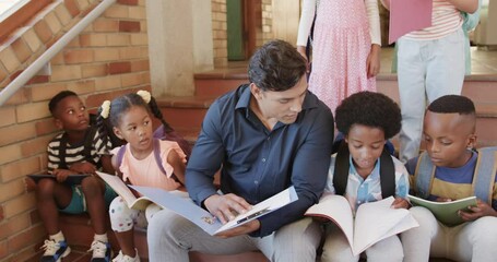 Diverse male teacher and children sitting on stairs reading books in elementary school, slow motion