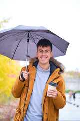Portrait of a smiling young man holding cup of coffee and umbrella in a rainy autumn day at the...