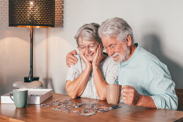 Carefree senior couple at home spend time together doing a puzzle on the wooden table. Elderly man and woman enjoying free time in retirement