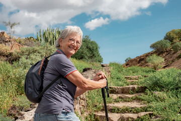Happy senior woman walking in a sunny day on trail outdoors on country excursion. Relaxed caucasian...