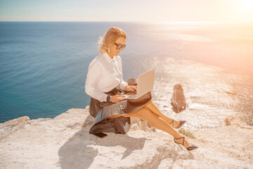 Business woman on nature in white shirt and black skirt. She works with an iPad in the open air with a beautiful view of the sea. The concept of remote work.