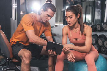 Healthy lifestyle and fitness concept. Asian woman wearing sports bra sitting on yoga ball looking at exercise schedule tablet with Caucasian male trainer in gym.