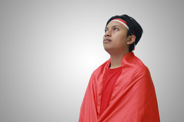 Portrait of thoughtful Asian man in t-shirt with red white ribbon on head, embraced by flag, celebrating Indonesia's independence day. Isolated image on gray
