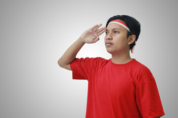 Portrait of attractive Asian man in t-shirt with red and white ribbon on head, giving salute pose with hand during Indonesian flag ceremony