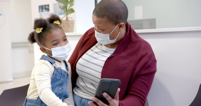 African american mother and daughter wearing face masks using smartphone in hospital, slow motion