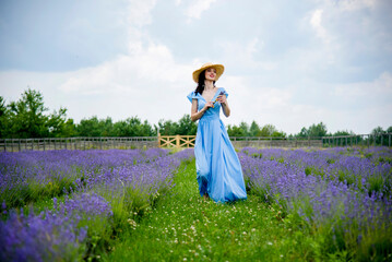 beautiful brunette smiling young woman in blue dress  with straw hat walking in lavender field on...