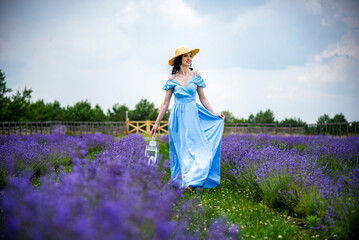 happy smiling beautiful brunette woman in a blue dress and straw hat in a lavender field holding wooden vintage white lamp in her hands