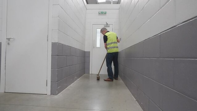 Male wearing yellow high vis vest brushes floor of building, locked off