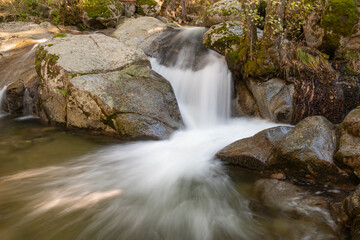 Fototapeta premium Small waterfall in a river whose flow runs between large granite stones deep in a dark forest. Concept of peace and relaxation, water stream and silk water.