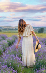 Caucasian girl in a light dress and hat walking in a lavender field, view from the back. Background sky and pink sunset. Provence, France. Travel concept.