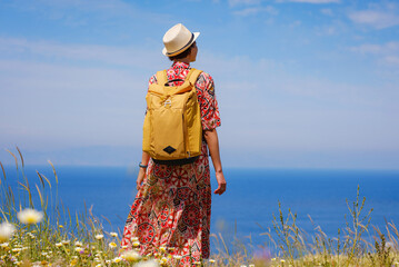 summer trip to Rhodes island, Greece. Young Asian woman in ethnic red dress and yellow backpack walk to viewpoint over city of Rhodes. Tourism, vacation, solo travel and discovery concept