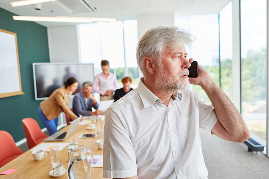 Bearded senior businessman talking on smart phone while sitting in front of colleagues discussing at office