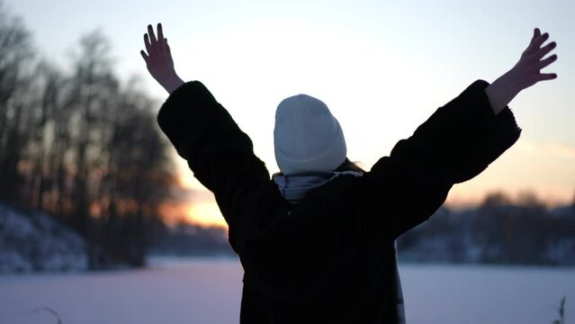 Back view unrecognizable excited woman stretching hands in slow motion admiring winter sunrise in dusk. Happy cheerful Caucasian lady enjoying weekend leisure at dawn outdoors