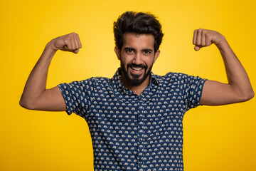 Fototapeta premium I am strong. Fit sporty indian man showing biceps and looking confident, feeling power strength to fight for rights, energy to gain success win. Hindu guy isolated on studio yellow background indoors