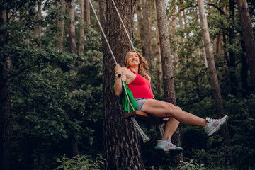 Beautiful pregnant woman having fun outside in the forest on a wooden swing swinging high up and enjoying nature