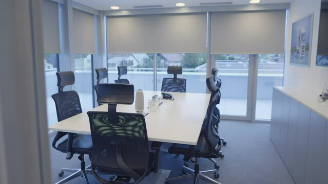 Office meeting room with no people view behind window with white blinds handheld shot. Desk and chairs in modern empty conference boardroom at day time