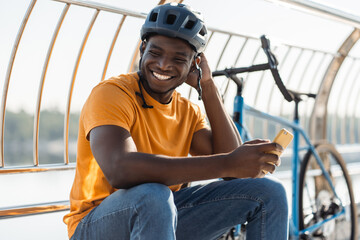 Authentic portrait of smiling African American man wearing protective helmet holding mobile phone,...