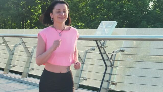 Young woman in pink top and dark leggings jogging across bridge near city park. Smiling lady with hands tattoo and neck accessory running on bridge