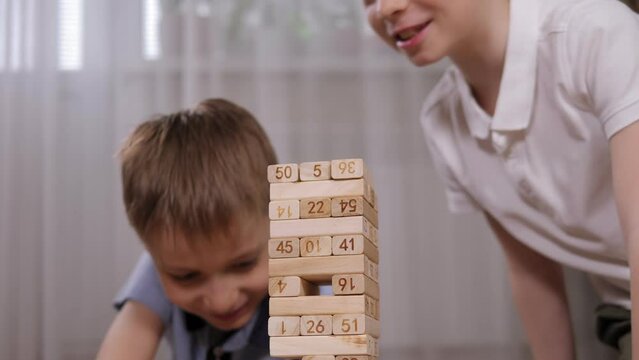 Two cheerful little boys are playing a board game with wooden blocks at home, they take out one wooden block and put it on top.