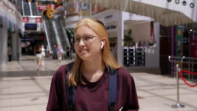 A stylish teenage girl in round glasses and wireless headphones with a backpack is standing in a shopping mall, with an escalator in the background.