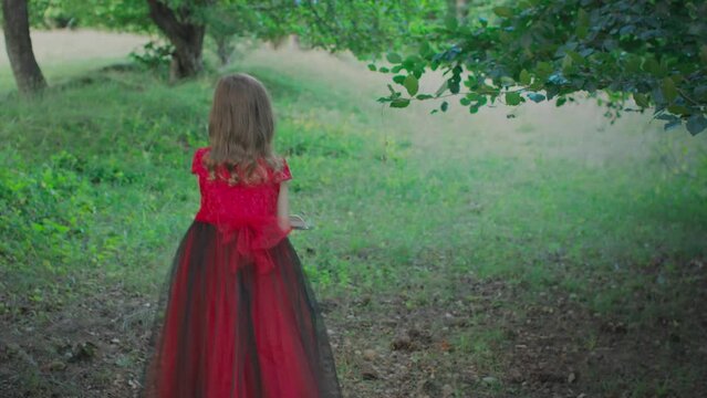 Little girl in red dress walking through the forest and reading a book. Concept of reading and imagination world