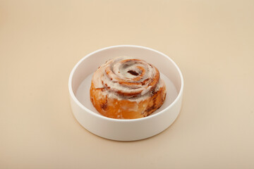 Freshly baked cinnamon bun or cinnabon roll in white plate on beige background. Selective focus, copy space