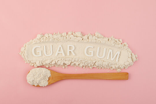 Guar gum powder or guaran in wooden spoon on pink background, top view. Pile of powder with Inscription GUAR GUM. Design element. Food additive E412. Fat Replacer, Dough Improver