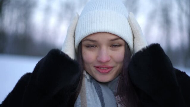 Front view portrait of happy carefree young woman touching hat in slow motion looking at camera with brown eyes and toothy smile. Positive charming Caucasian lady posing in morning winter forest