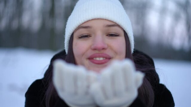 Headshot of cheerful woman blowing white snow from palms in slow motion looking at camera. Front view closeup of joyful happy relaxed Caucasian lady posing in winter forest outdoors