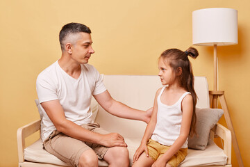 Fototapeta premium Young handsome father talking with his daughter sitting on cough at home against beige wall having conversation about their relationship looking at each other.