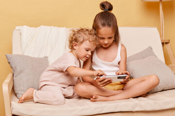 Concentrated children little girls sisters sitting on couch using smartphone together posing against beige wall playing on mobile phone looking at device display.