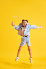 Full-length portrait of active little girl, child in casual clothes cheerfully jumping against yellow studio background. Concept of emotions, childhood, education, fashion, lifestyle, ad