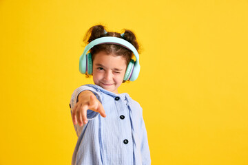 Portrait of positive little girl child listening to music in headphones and grimacing against yellow studio background. Concept of emotions, childhood, education, fashion, lifestyle, ad