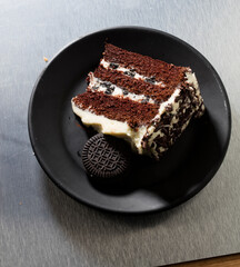 Tarta de chocolate con crema y cookie sobre plato negro y fondo blanco, vista cenital. Chocolate cake with cream and cookie on black plate and white background, overhead view.